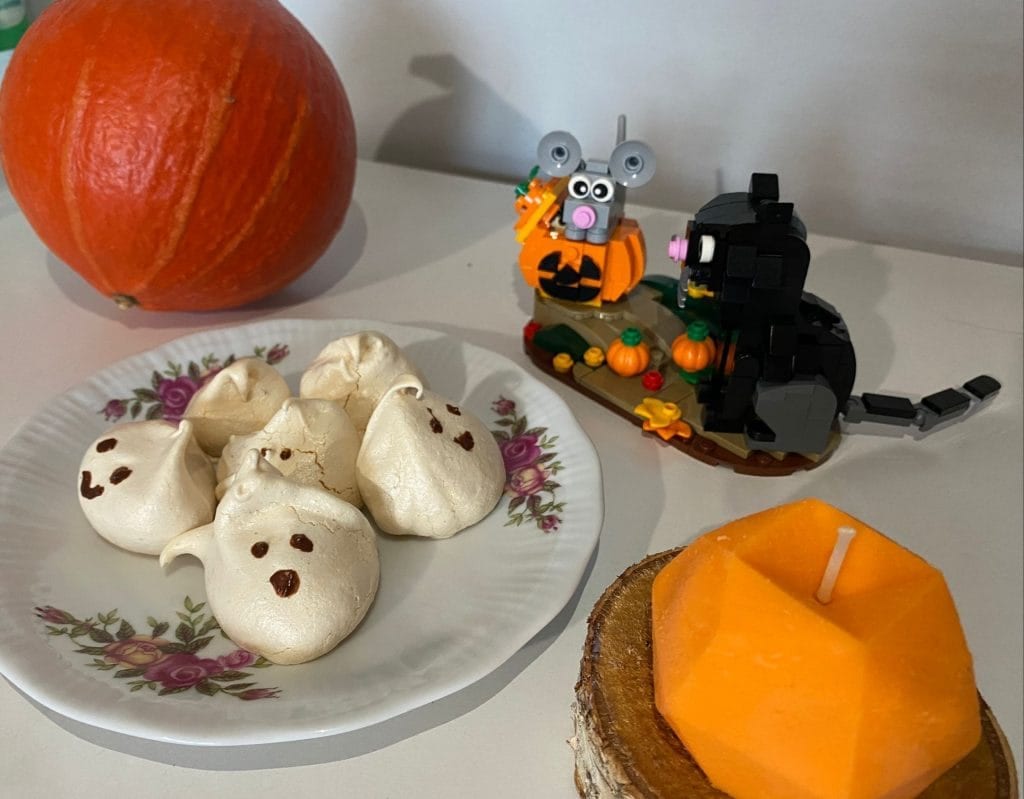 Talerz z bezami, które mają domalowane czekoladowe buźki i oczka, obok dynia hokkaido, halloweenowy zestaw z Lego i pomarańczowa świeczka.
