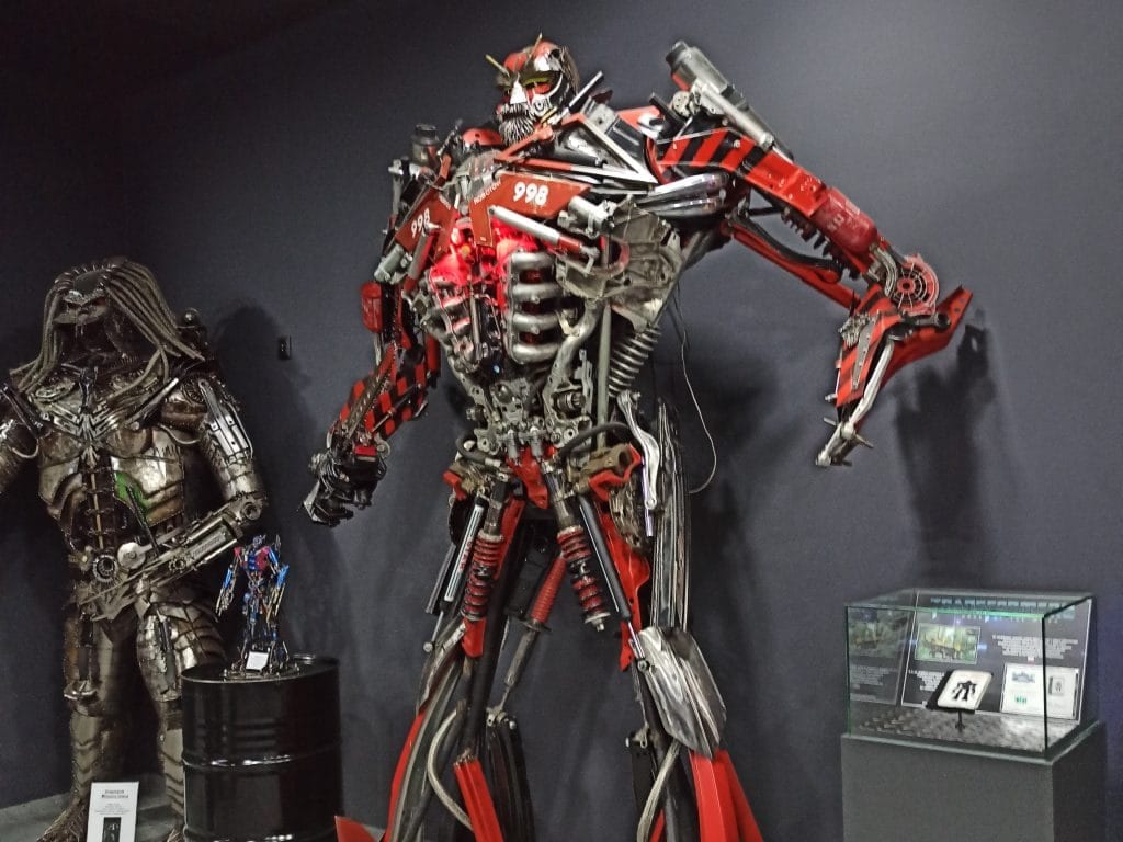 Na pierwszym planie potężny czerwony robot wykonany ze złomu. Na drugim planie rzeźba przedstawiająca Predatora.